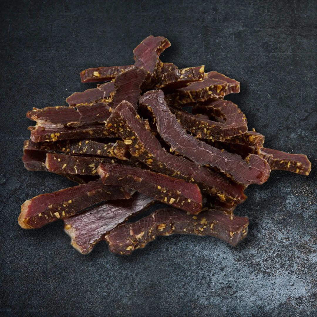 A close-up of air-dried biltong slices with visible spices on a dark textured background