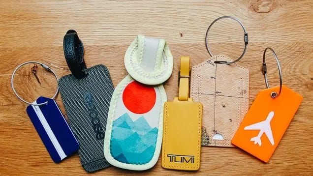 A collection of five unique luggage tags displayed on a wooden surface, featuring various designs and colors, including blue, yellow, orange, and patterned styles