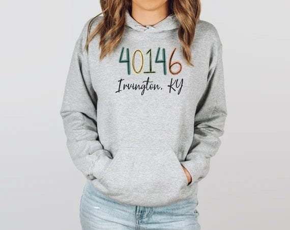 A woman wearing a gray hoodie featuring the zip code "40146" in colorful, stitched-style numbers with "Irvington, KY" written below in cursive font