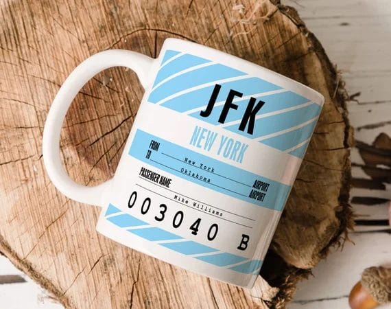 A white ceramic mug featuring a JFK New York airport ticket design with blue diagonal stripes, including passenger details and flight information for a personalized touch.