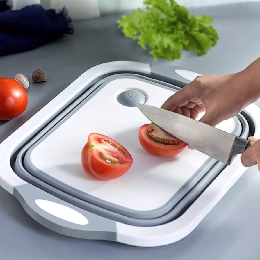 A person cutting a tomato on a modern, multi-functional cutting board. The board is white with gray edges and features a collapsible design, which can also function as a wash basin. Fresh vegetables, including tomatoes and lettuce, are placed nearby on the countertop.