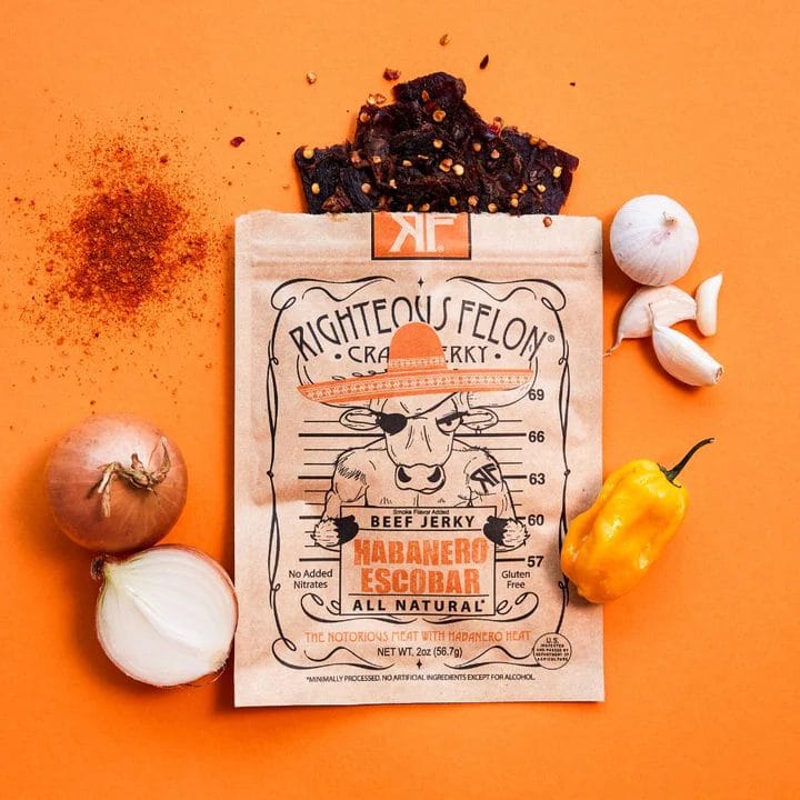 Righteous Felon Habanero Escobar Craft Jerky in unique packaging, surrounded by habanero pepper, onion, garlic, and chili powder on an orange background.
