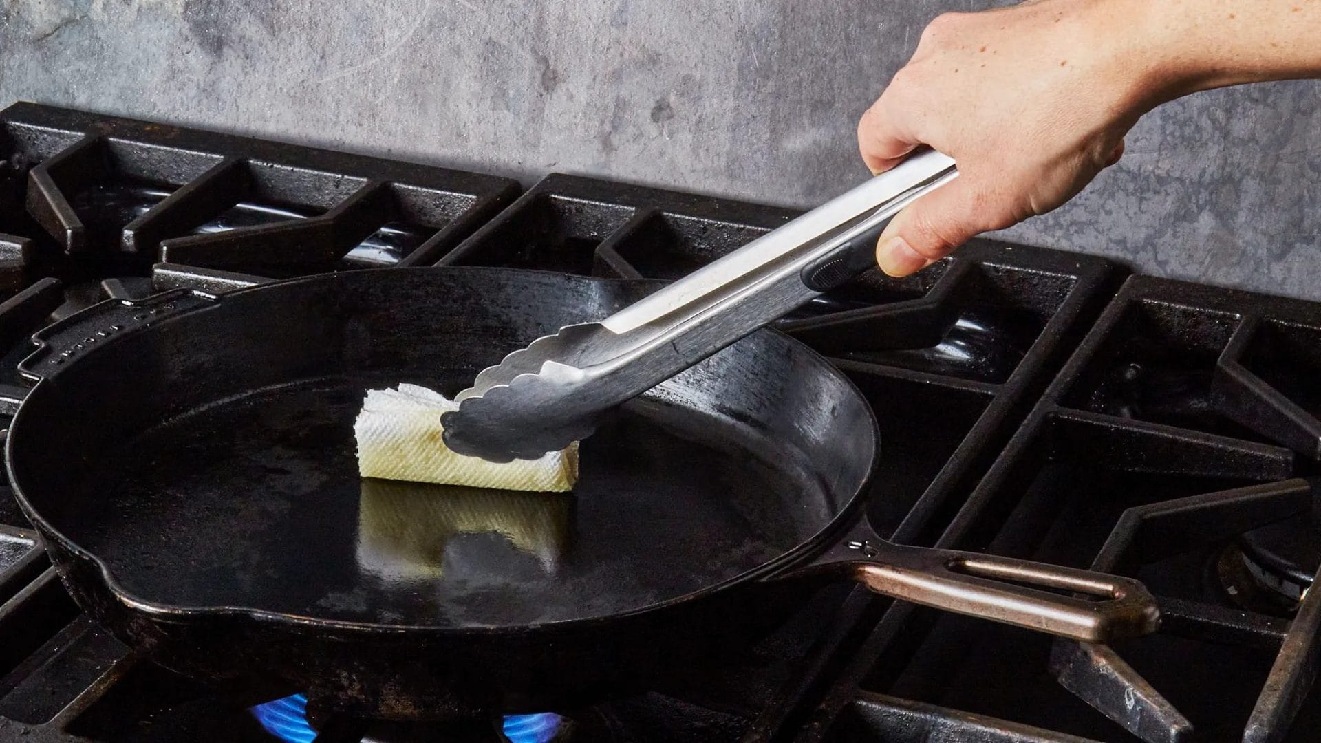 A person seasoning a cast iron skillet on a gas stove using tongs and a paper towel soaked in oil. 