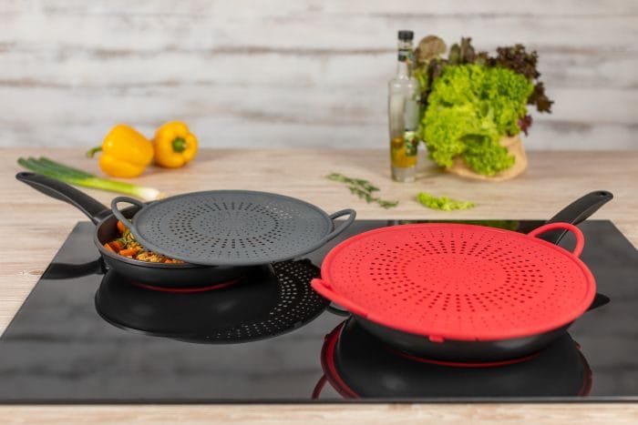 Two silicone splatter screens (one red, one black) placed on frying pans on a stovetop. These covers prevent oil splatters while allowing steam to escape, keeping the kitchen clean and reducing mess during cooking.