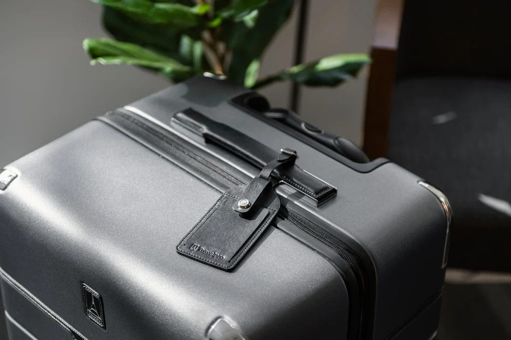  A modern, dark grey hard-shell suitcase with a black leather luggage tag attached to the handle, placed indoors with a potted plant in the background