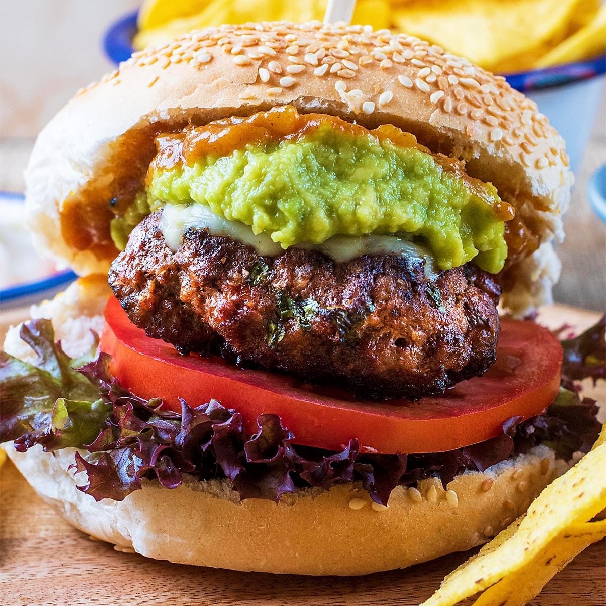 Close-up of a spicy guacamole burger featuring a juicy beef patty with herbs, melted cheese, fresh guacamole, tomato slices, and leafy lettuce on a sesame seed bun, served with tortilla chips.