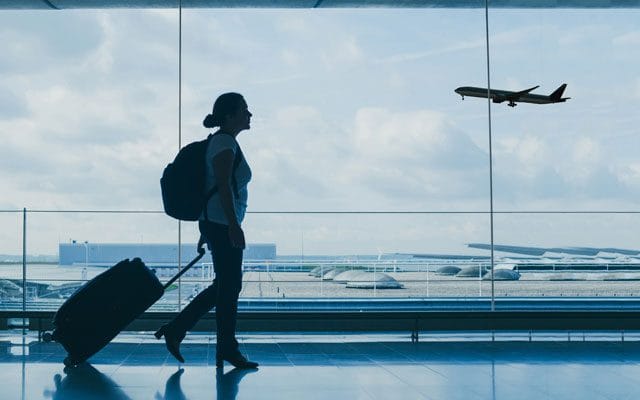 A woman carrying a backpack and pulling a suitcase walks past a large window at the airport, with an airplane taking off in the background