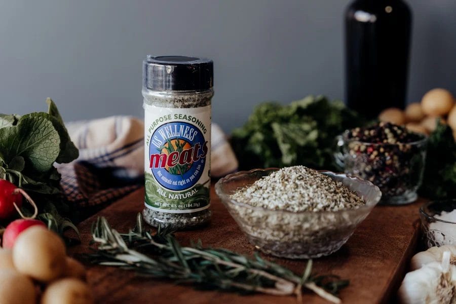 A bottle of US Wellness Meats All-Purpose Seasoning on a wooden table with herbs and dried spices