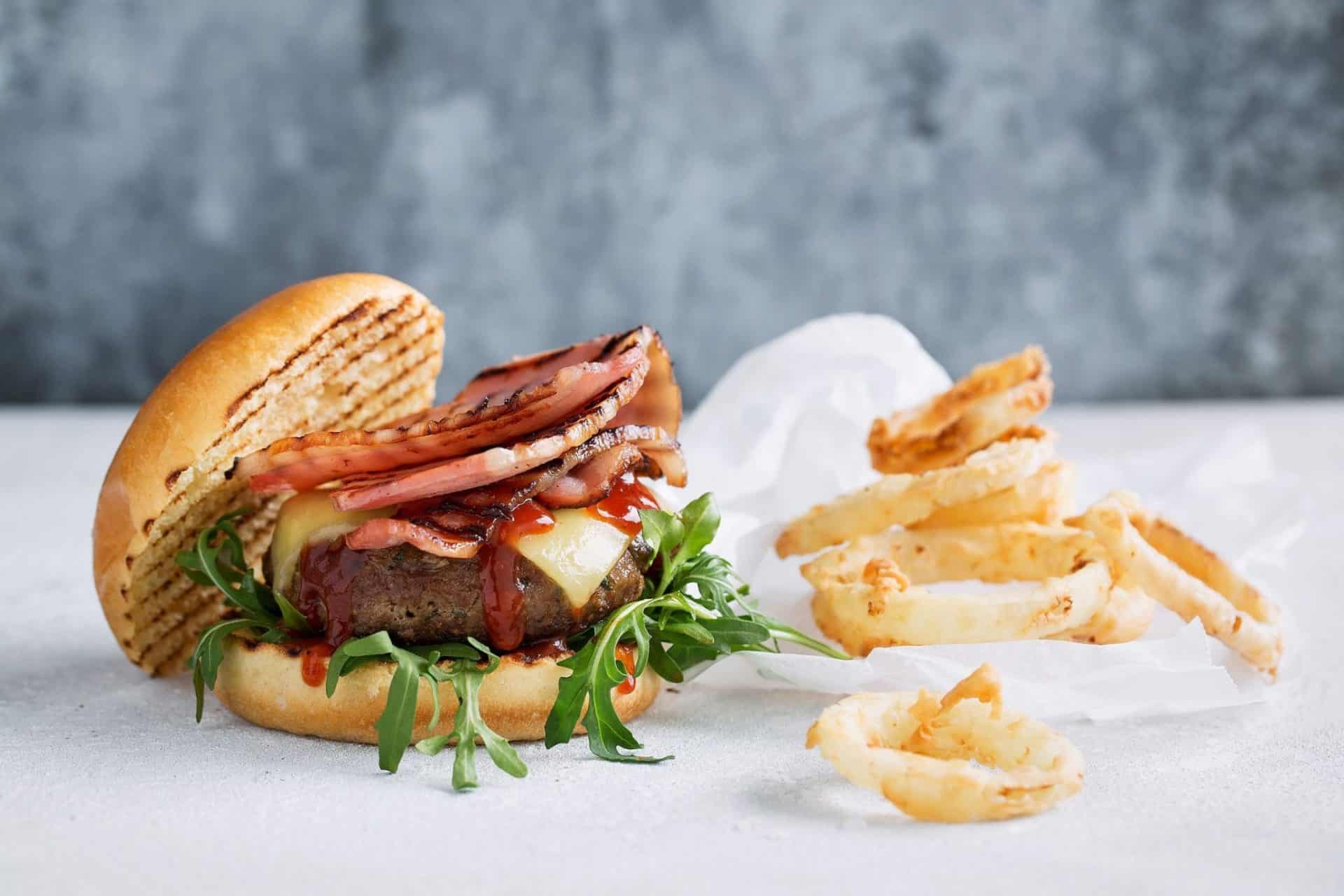 A gourmet BBQ bacon cheeseburger with melted cheese, crispy bacon strips, fresh arugula, and tangy barbecue sauce on a toasted bun, served with a side of golden onion rings.